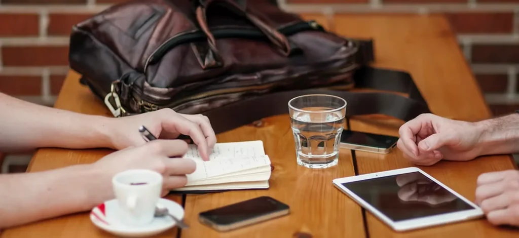 Two people meeting at a wooden table with notebooks, a tablet and coffee