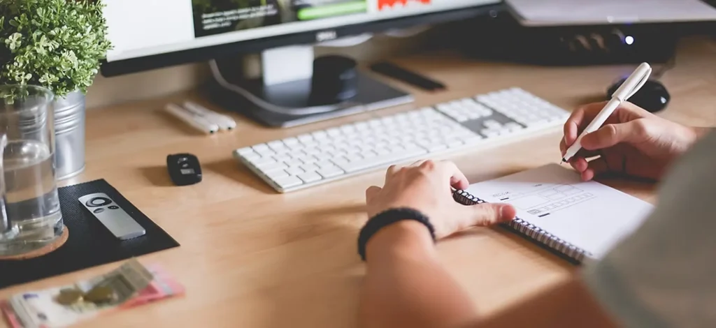 WEBSITE-DESIGN-service Hands sketching in a notebook at a desk with a keyboard and monitor